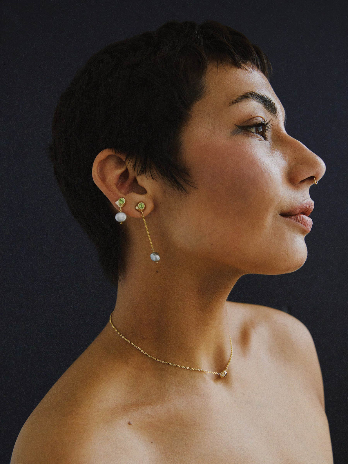 FARIS KIRA Drops in 14k gold-plated bronze with peridot, both sides of the pair styled on models right ear. Long side worn on standard lobe piercing, short side of pair worn on upper lobe ear piercing. Styled with KIRA Necklace in 14k gold with garnet