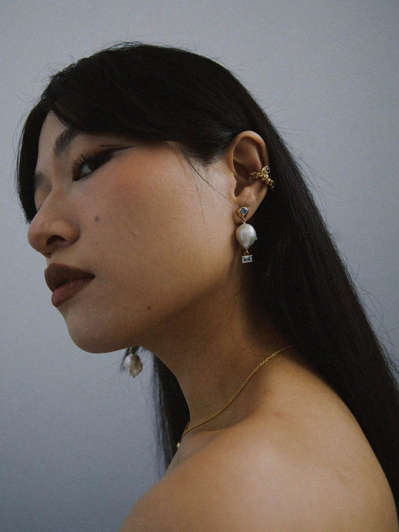 FARIS VENUS Drops in gold-plated bronze with lab-created blue sapphire, and topaz, shown on model. Styled with ROCA GEM Ear Cuff in gold-plated bronze with garnet. Short side of asymmetrical earring shown